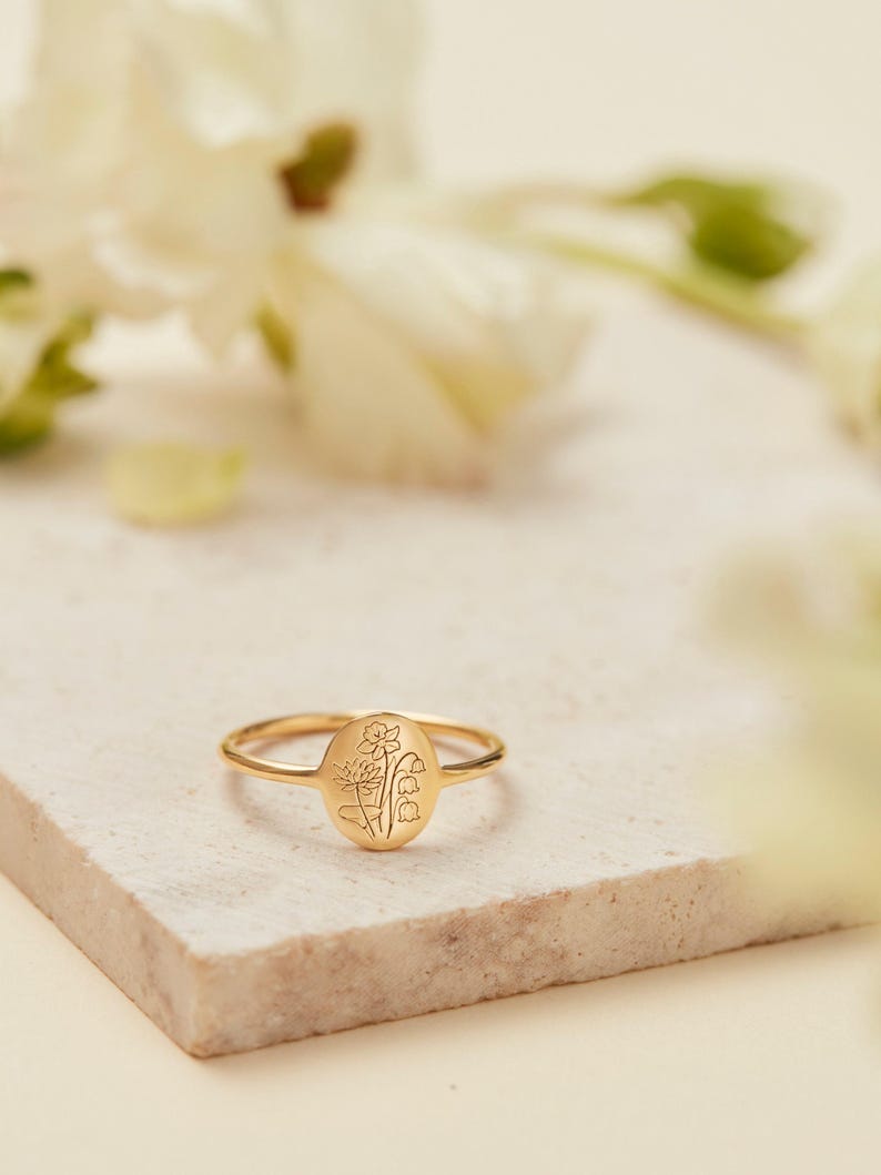 May include: A gold-colored ring with an oval face featuring an engraved floral design. The ring is set on a light-colored, textured surface, with blurred white flowers in the background.
