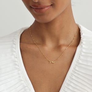 May include: A fine gold necklace with a script pendant. The necklace is worn around the neck and rests on a white top. The pendant features Hebrew script. The chain is thin and appears to be made of gold.