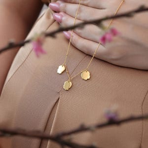 May include: A gold-colored necklace with three pendant charms. The charms are in the shape of a heart, a profile, and a rounded shape. The necklace is displayed on a beige background, with a blurred floral branch in the foreground.