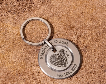 Puede incluir: Un llavero plateado con un colgante redondo que presenta un diseño de huella dactilar. El llavero está grabado con los nombres "Stephanie" y "Josh", un símbolo de corazón y la fecha "14 de febrero de 2022".