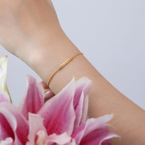 May include: A delicate gold bracelet with a small rectangular bar engraved with the names "GRACE" and "BRIAN". The bracelet features a fine chain and small gemstones. The bracelet is worn on a wrist.