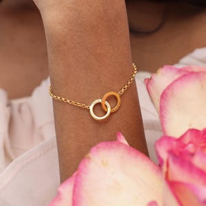 May include: A gold-colored bracelet featuring two interlocking rings, each inscribed with a name. The bracelet is connected by a delicate chain. The image is set against a soft background with pink rose petals.