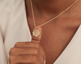 May include: A gold-colored necklace with a circular pendant. The pendant features a detailed fingerprint design. The necklace is worn against a white shirt. The chain is delicate and the pendant is held up by a finger.