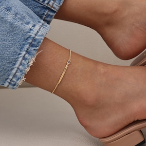 May include: A delicate gold anklet with a small rectangular blue gemstone and engraved text. The anklet is worn on an ankle, with a denim-clad leg and a sandal visible in the background. The anklet is a simple, elegant piece of jewelry.
