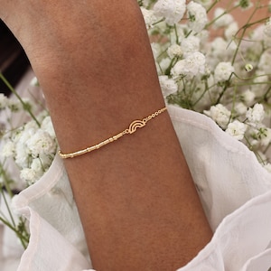 May include: A delicate gold bracelet featuring a rainbow charm and a series of small beads. The bracelet is worn on a wrist, with a background of white flowers and a white fabric.
