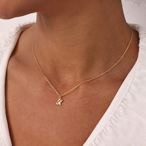 May include: A delicate gold-colored necklace featuring a small cursive letter "L" pendant. The pendant hangs from a thin chain, creating a minimalist and elegant look. The necklace is worn against a white top.