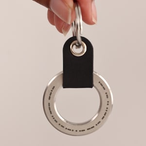 May include: A silver metal key ring with a black leather strap. The ring has a series of dotted and dashed markings around its circumference. The key ring is held by a hand.