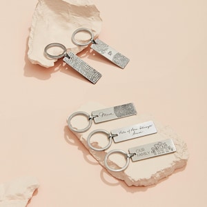 May include: Silver keychains with fingerprint and text designs. One keychain has the text "&", another has a fingerprint, and others have the words "Mom", "Lots of love always", and "OUR FAMILY" with a family illustration.