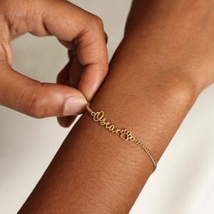 May include: A delicate gold-colored bracelet with the name "Oscar" in cursive script and a paw print charm. The bracelet is being clasped on a wrist, showcasing its fine chain and personalized design.