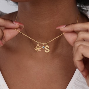 May include: A gold necklace featuring a flower charm, a small blue gem, and the letter "S". The necklace is held up by a person. The chain is delicate, and the charms are small and detailed.