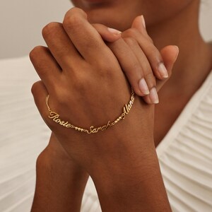 May include: A gold-colored bracelet with the names "Rosie", "Sarah", and "Alana" inscribed in a delicate script. The bracelet features small, round beads between the names. The bracelet is displayed on a person's wrist.