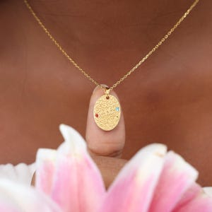 May include: A gold necklace featuring an oval pendant with a textured pattern. The pendant has a red and a blue gemstone, and the engraved text "Lulu & Mia". The necklace is set against a blurred pink flower background.