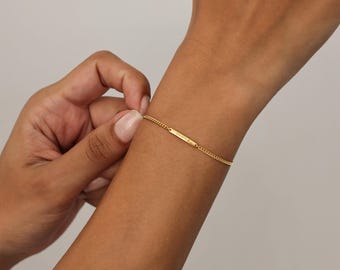 Peut inclure: Un bracelet en or fin avec un pendentif rectangulaire. Le bracelet est porté sur un poignet, et le pendentif porte un texte gravé. La chaîne est de style gourmette fine. Le fond est de couleur neutre.