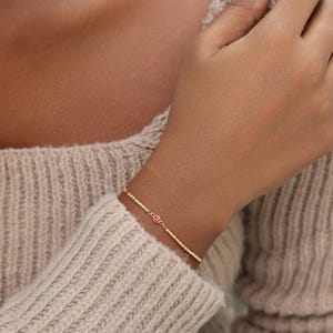 May include: A delicate gold chain bracelet featuring a series of small, evenly spaced gold beads along the band and a single, round, vibrant pink gemstone set in a gold bezel at the center of the design, creating a refined and minimalist look.