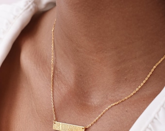 May include: A gold-colored necklace with a rectangular pendant featuring a fingerprint design. The pendant hangs from a delicate chain. The necklace is worn around the neck.