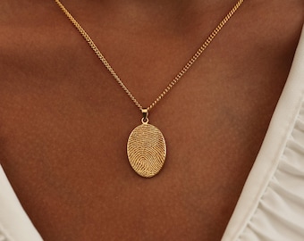 Puede incluir: Collar con colgante de huella dactilar dorado. El colgante ovalado presenta un diseño detallado de huella dactilar. El collar tiene una cadena delicada y se lleva alrededor del cuello. La joya es un recuerdo personalizado.