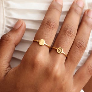 May include: Two gold-colored rings. One ring features a circular design with a pattern, and the other ring has a paw print design. The rings are on a person's finger.