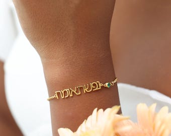 May include: A gold-colored bracelet with Hebrew text is worn on a wrist. The bracelet has a small green gemstone. The background includes a white garment and orange flowers.