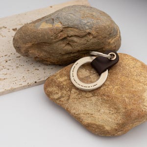 May include: A silver-colored metal keychain with a dark brown leather strap. The keychain has a circular design with engraved dots and dashes, likely Morse code. The keychain is resting on a large, textured rock.