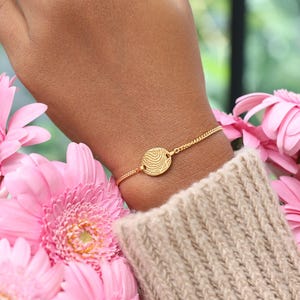 May include: A gold-toned bracelet featuring a small, circular charm engraved with a detailed fingerprint pattern. The charm is centered on a delicate, fine-link chain that rests against the skin, providing a subtle and personalized accessory.
