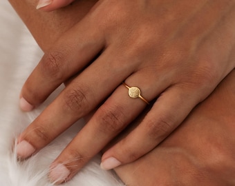 May include: A gold ring featuring a small, textured, circular design. The ring is on a thin band and is worn on a finger. The ring is set against a soft, white background.