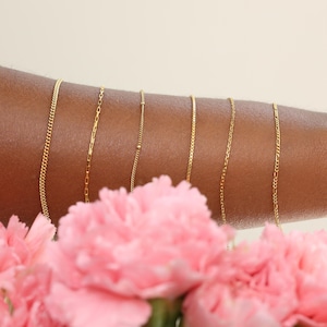 May include: Six distinct gold-toned metal chains of varying link styles, including curb, cable, and figaro patterns, arranged vertically in a row. The chains are displayed against a skin-toned surface, partially obscured by soft pink flowers.