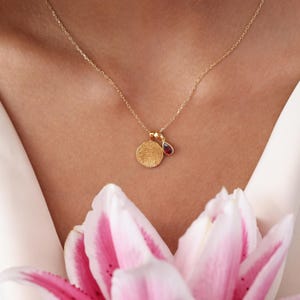 May include: A delicate gold necklace featuring a fingerprint pendant and a small, teardrop-shaped red gemstone charm. The necklace is displayed against a soft, neutral background, with a hint of pink and white flowers in the foreground.