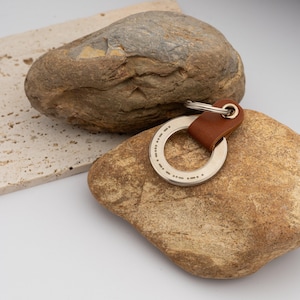 May include: A silver-colored keychain with a brown leather strap. The circular metal piece has a series of dots and dashes engraved around the edge. The keychain is resting on a brown rock, with another rock and a light-colored stone slab in the background.