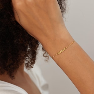 May include: A delicate gold bracelet featuring a rectangular bar pendant. The bracelet is attached to a fine chain with small, round beads. The jewelry is worn on a wrist, with a white shirt visible in the background.