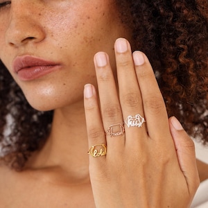 May include: Three personalized name rings in gold, rose gold, and silver. The rings spell out the names "Lola", "Bella", and "Abby" in a delicate, cursive font. The rings are displayed on a hand with a light skin tone.
