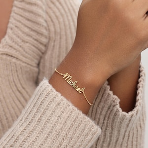 May include: A gold-toned chain bracelet featuring a central nameplate with the name Michael written in a cursive script font. The delicate metal chain connects to the nameplate on both sides, creating a continuous piece of jewellery.