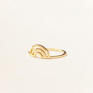 May include: A gold-colored ring featuring a rainbow design. The ring has a delicate, minimalist style, with the rainbow shape as the focal point. The ring is set against a white background.