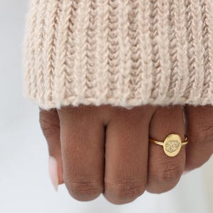 May include: A gold ring featuring a small, oval-shaped face with a textured, granular surface. The band is thin and circular, maintaining a consistent metallic finish throughout the design. The ring is worn on the ring finger of a hand.