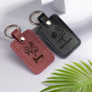 May include: Two leather keychains with silver keyrings. One is burgundy with a floral design and the name "Jenny" engraved. The other is dark gray with a floral design and the text "Mama's garden".