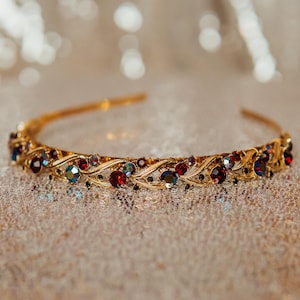 May include: A gold-coloured tiara with a delicate design. The tiara is decorated with red, blue, and iridescent gemstones. The gemstones are set in a floral pattern. A beautiful accessory for special occasions.