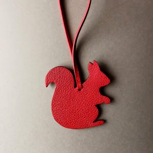 Könnte beinhalten: Ein roter, lederner, eichhörnchenförmiger Anhänger mit einer Schlaufe zum Aufhängen. Das Eichhörnchen hat eine strukturierte Oberfläche und ist von leuchtend roter Farbe. Der Hintergrund ist ein neutrales Grau.