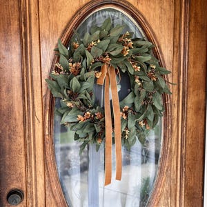 May include: A circular wreath composed of dark green leaves and small clusters of light brown foliage, accented with a long, vertical, solid tan ribbon hanging from the top center, mounted on an oval glass panel within a wooden door frame.