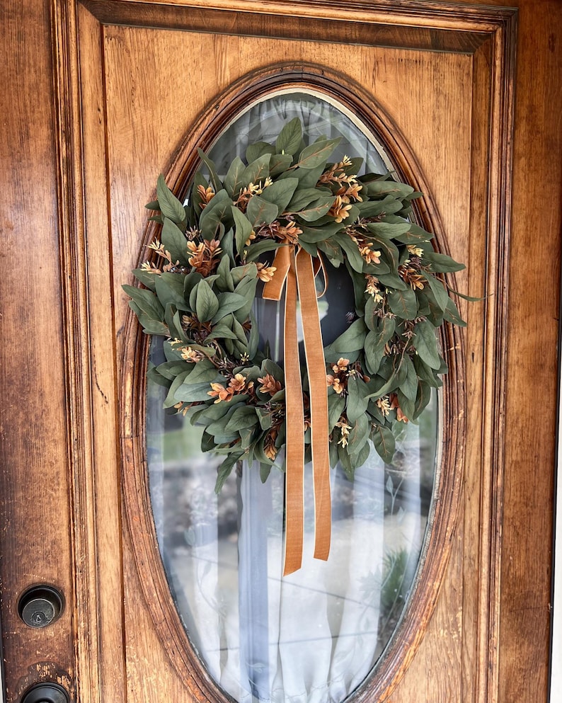 May include: A circular wreath composed of dark green leaves and small clusters of light brown foliage, accented with a long, vertical, solid tan ribbon hanging from the top center, mounted on an oval glass panel within a wooden door frame.