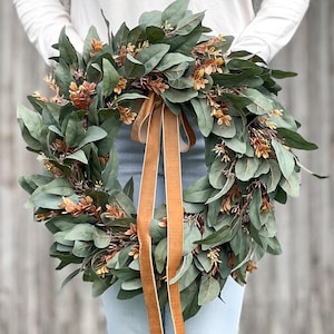 May include: A circular wreath composed of dense dark green leaves and small clusters of light brown foliage. Two long, smooth, tan-colored velvet ribbons hang vertically from the center of the wreath, extending downward from the top inner edge.