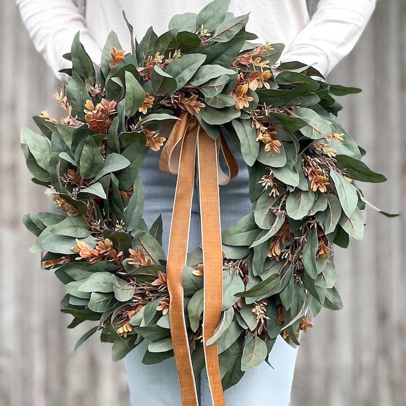 May include: A circular wreath composed of dense dark green leaves and small clusters of light brown foliage. Two long, smooth, tan-colored velvet ribbons hang vertically from the center of the wreath, extending downward from the top inner edge.