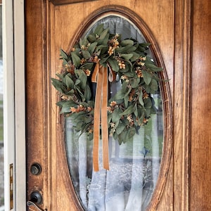 May include: A circular wreath featuring dark green eucalyptus leaves and small brown floral accents, adorned with a long, hanging tan velvet ribbon. The wreath is mounted on the oval glass window of a wooden door with a metal handle and lock.