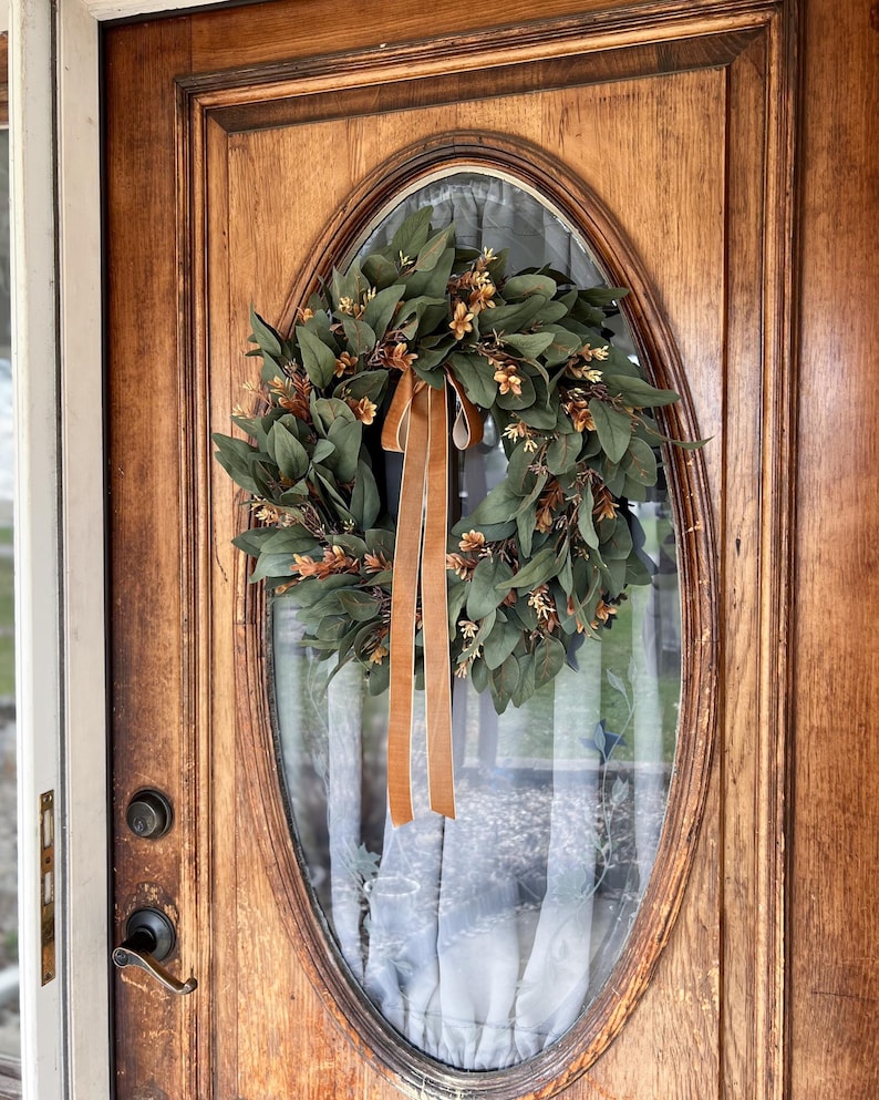 May include: A circular wreath featuring dark green eucalyptus leaves and small brown floral accents, adorned with a long, hanging tan velvet ribbon. The wreath is mounted on the oval glass window of a wooden door with a metal handle and lock.