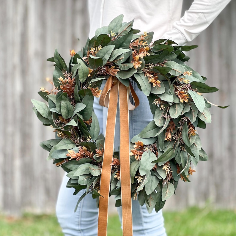 May include: A circular wreath composed of dark green eucalyptus leaves and small clusters of copper-colored foliage. Two long, smooth, tan-colored velvet ribbons hang vertically from the center of the wreath, extending downward.