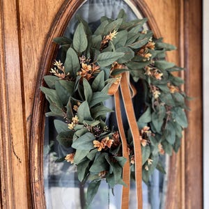 May include: A circular wreath composed of dark green eucalyptus-style leaves and small clusters of light brown, dried-looking flowers. Two long, smooth, tan-colored ribbons hang vertically from the center of the wreath, adding a soft texture.