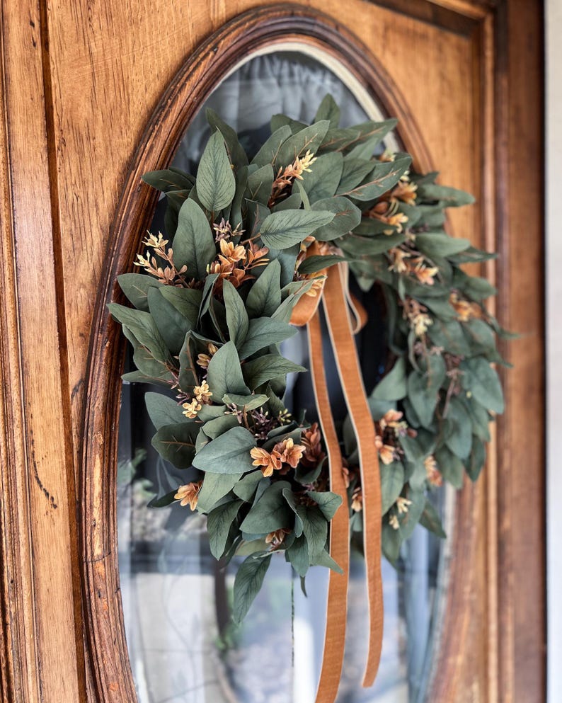 May include: A circular wreath composed of dark green eucalyptus-style leaves and small clusters of light brown, dried-looking flowers. Two long, smooth, tan-colored ribbons hang vertically from the center of the wreath, adding a soft texture.