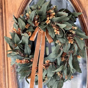 May include: A circular wreath featuring dark green eucalyptus-style leaves interspersed with small, light brown floral sprigs. A long, smooth, tan-colored velvet ribbon is tied at the top, with two long tails hanging down through the center of the wreath.