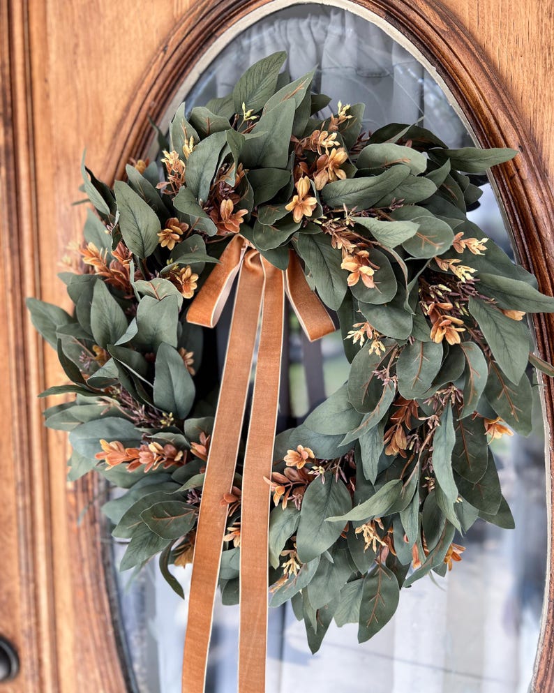 May include: A circular wreath featuring dark green eucalyptus-style leaves interspersed with small, light brown floral sprigs. A long, smooth, tan-colored velvet ribbon is tied at the top, with two long tails hanging down through the center of the wreath.