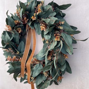 May include: A circular wreath featuring dense, dark green oval leaves interspersed with small, light brown floral clusters. A smooth, tan-colored ribbon hangs from the top, extending downward through the center of the foliage arrangement.