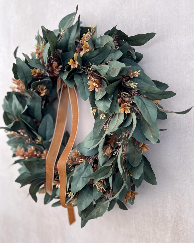 May include: A circular wreath featuring dense, dark green oval leaves interspersed with small, light brown floral clusters. A smooth, tan-colored ribbon hangs from the top, extending downward through the center of the foliage arrangement.