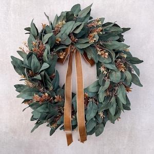 May include: A circular wreath constructed from dark green eucalyptus leaves and interspersed with small brown sprigs. A long, smooth brown velvet ribbon is tied at the top center, with two tails hanging down through the middle of the wreath.